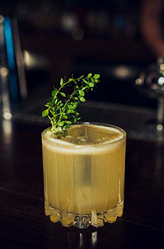 Yellow Orangano whiskey cocktail in glass on bar with oregano sprig garnish - Redemption Whiskey