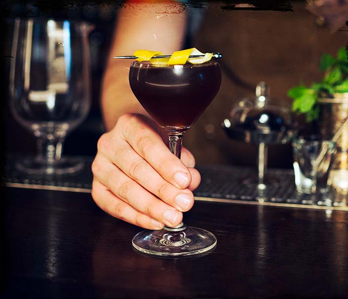 bartender serving dark brown better late than never whiskey cocktail with lemon twist