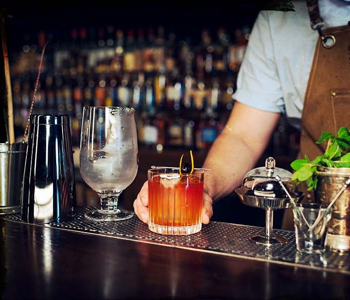 bartender serving red brown labyrinth whiskey cocktail with orange peel and cherry garnish at bar